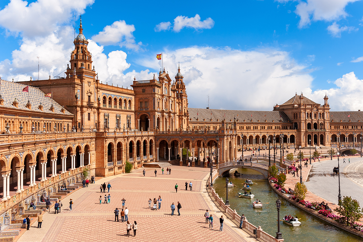 Wide view of Plaza de España in Seville, Spain, featuring a grand semicircular brick building with ornate towers, a canal crossed by decorative bridges, a large patterned plaza with a central fountain, and visitors walking throughout the historic landmark under a cloudy sky.