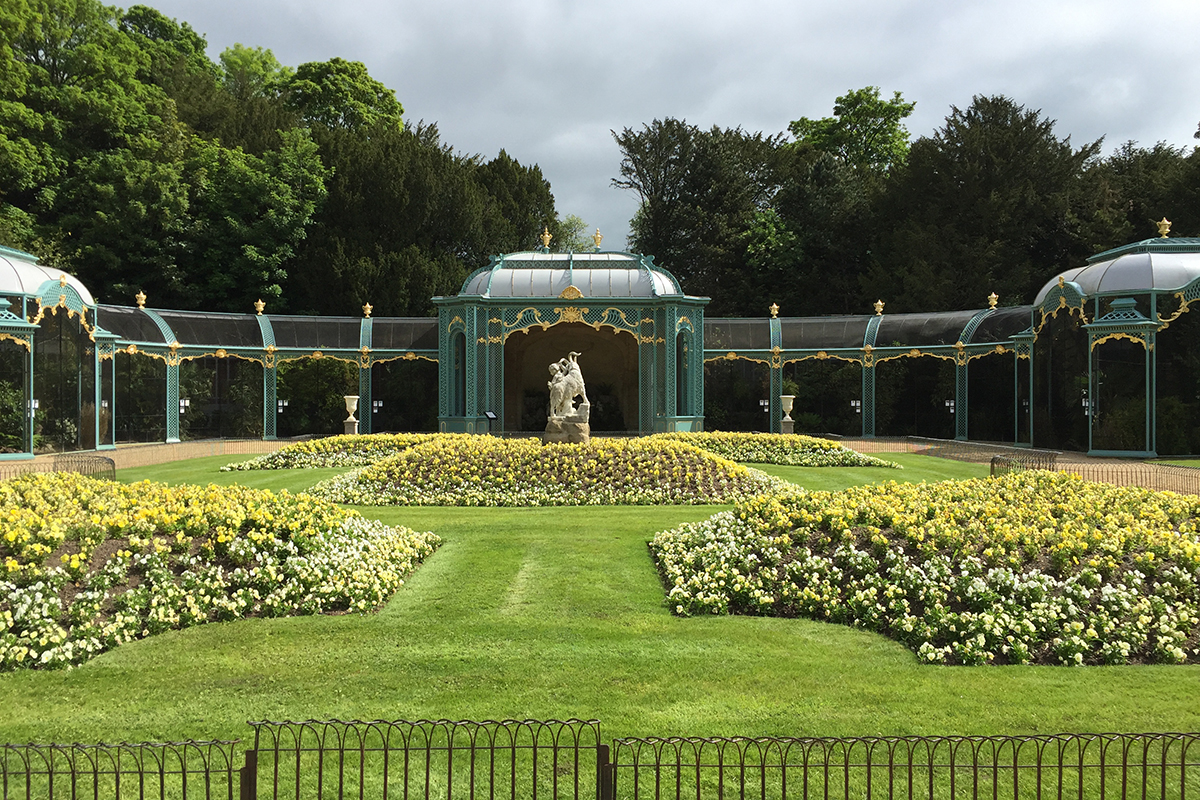 Formal garden with neatly trimmed lawns and large beds of yellow and white flowers leading to a central stone sculpture. An ornate green-and-gold glass aviary curves around the garden, with dense trees and a cloudy sky in the background.