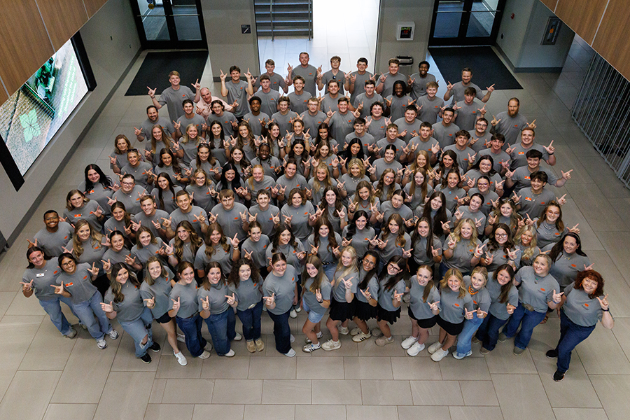 Overhead group photo of more than 100 students and staff gathered in a large, modern building lobby. Participants wear matching gray T-shirts and face the camera while making the OSU hand sign.